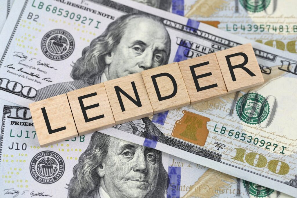 Wooden blocks spelling ‘LENDER’ placed on U.S. hundred-dollar bills, representing peer lending versus licensed moneylender risks and benefits.